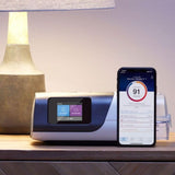 Smartphone displaying sleep tracking app next to CPAP machine on a bedside table with a lamp.