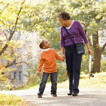 A grandmother and grandson walking hand in hand through a park, smiling and enjoying a sunny day, with yellow autumn leaves in the background.