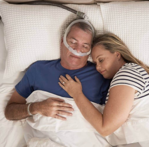 Man wearing a CPAP machine sleeping in bed next to a woman, showcasing sleep apnea treatment and companionship.