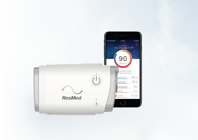 ResMed AirMini CPAP device next to a smartphone displaying sleep score app interface, against a sky background.