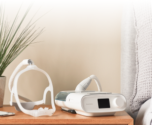 CPAP machine and mask on a wooden nightstand beside a bed with a gray blanket, plant in the background.