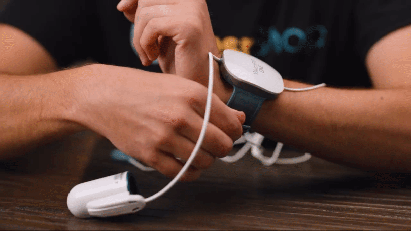A person assembles a medical device with cables that attaches to the wrist, placed on a wooden table.